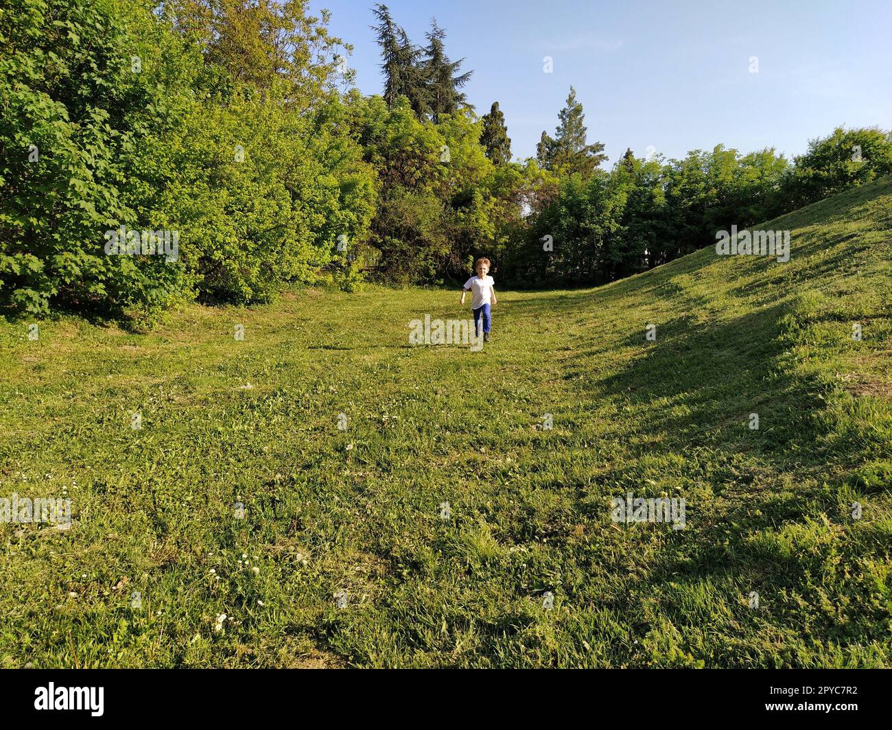 Un bambino corre nel parco. Un ragazzo con una T-shirt bianca e pantaloni blu. Prato o campo verde. Prato falciato. Cammina e gioca nella foresta. Attività sportive attive. Foto Stock