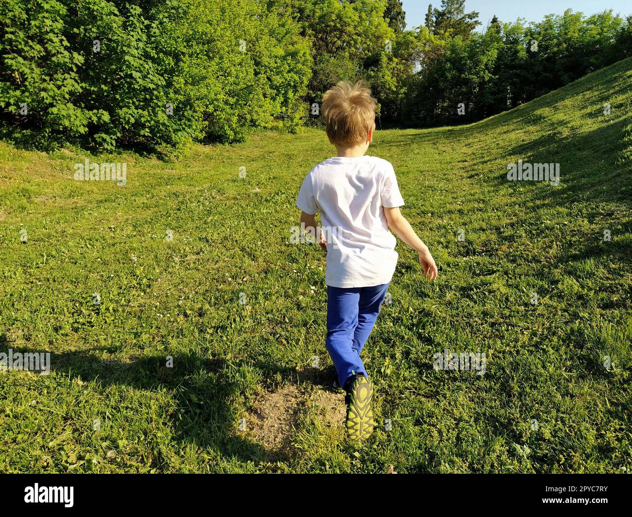 Un bambino corre nel parco. Un ragazzo con una T-shirt bianca e pantaloni blu. Prato o campo verde. Prato falciato. Cammina e gioca nella foresta. Attività sportive attive. Il ragazzo ha voltato le spalle alla telecamera Foto Stock