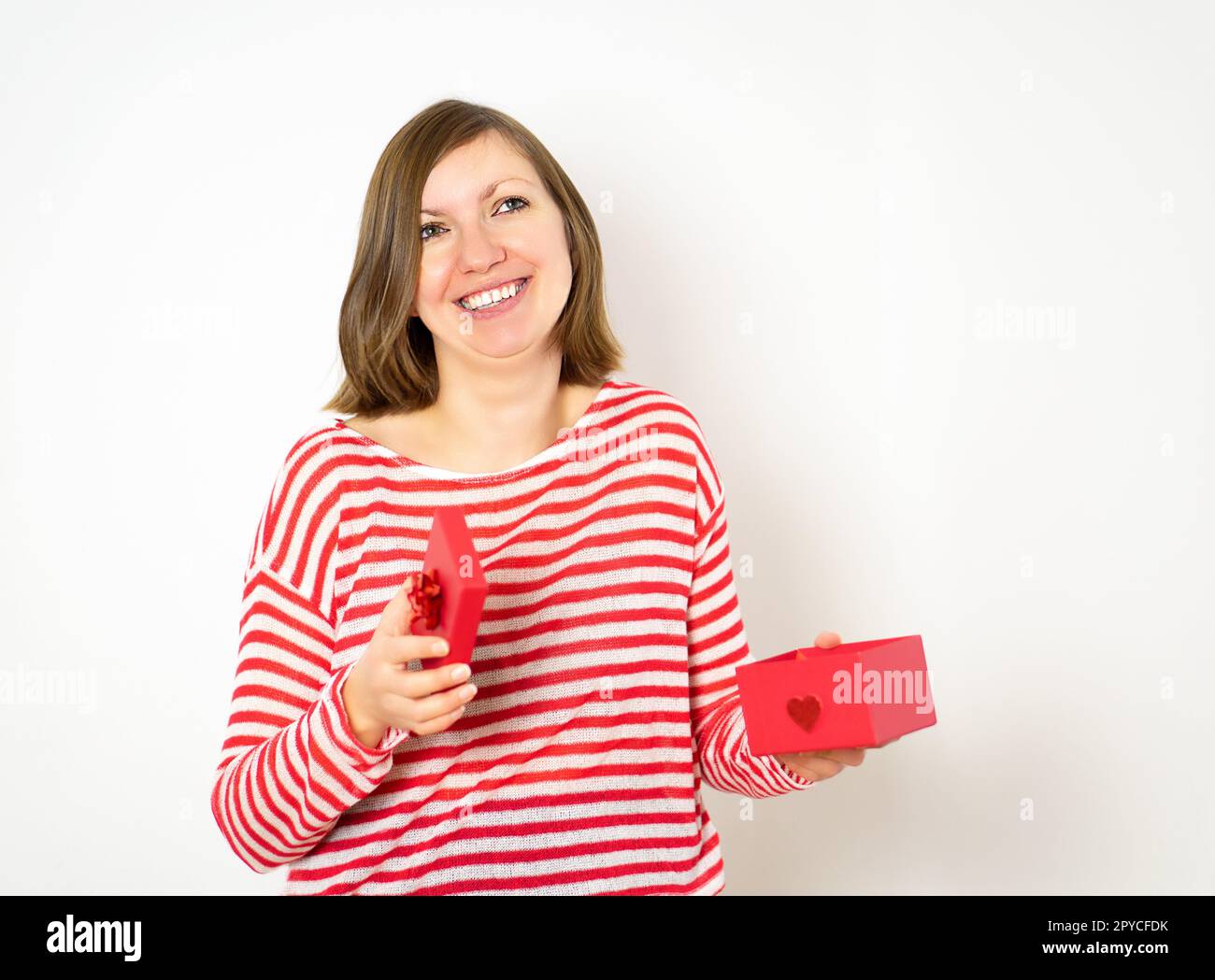 Ritratto di una bella ragazza sorridente felice in camicia rossa e bianca che apre una scatola regalo rossa su sfondo bianco. San Valentino concetto. Foto Stock