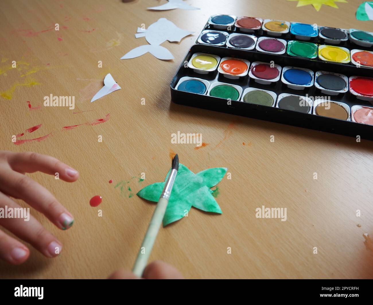 Desktop con strumenti di disegno. Set di vernici ad acquerello. Figurine di pesci dipinte tagliate a carta. Vista dall'alto. Lezione o attività creativa. La mano di un bambino con un pennello dipinge una stella Foto Stock