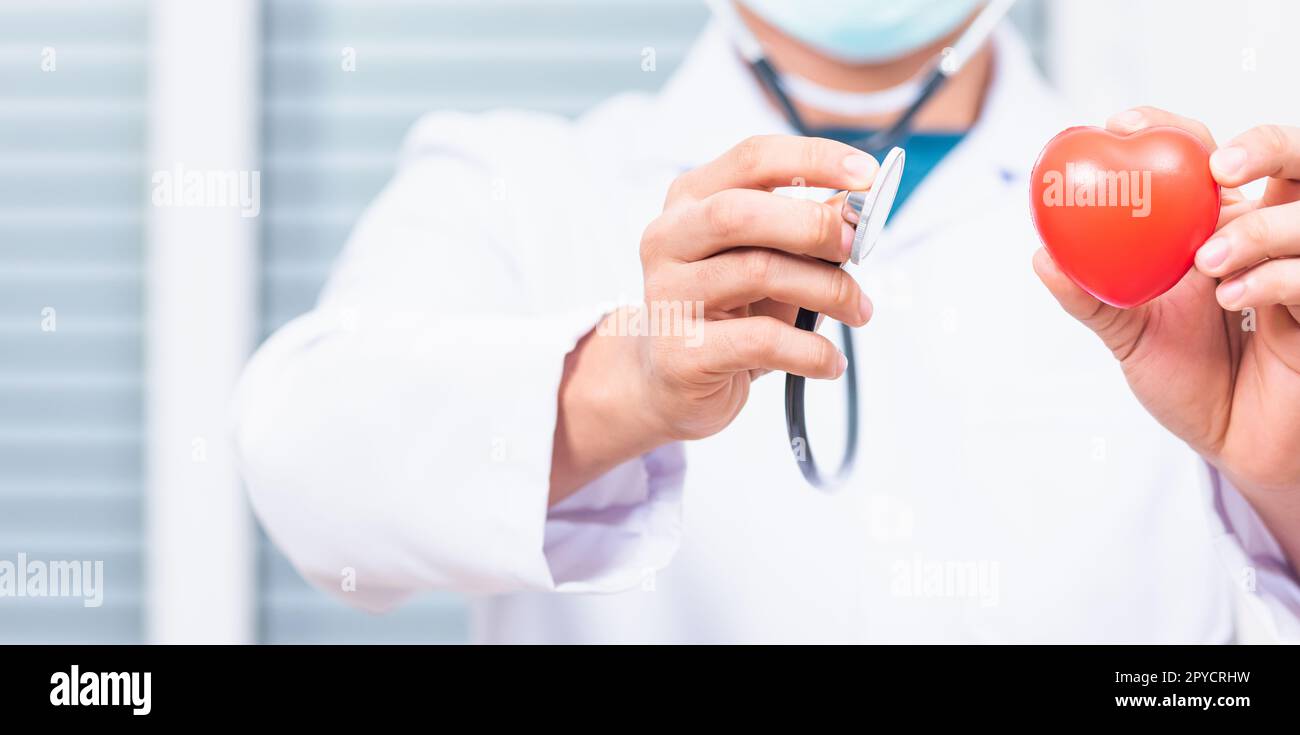 un medico che indossa un cappotto bianco tiene il suo stetoscopio a portata di mano per ascoltare esaminare il cuore rosso Foto Stock