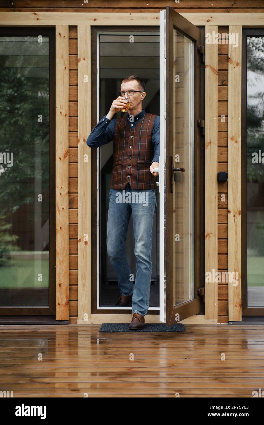 L'uomo di mezza età esce sulla terrazza di una piccola casa di legno con un vetro in mano Foto Stock L'uomo di mezza età esce sulla terrazza di una piccola casa di legno con un vetro in mano Foto Stock