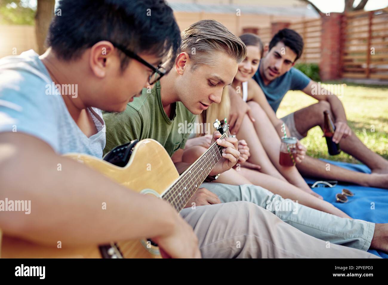 Strimpellimento via durante l'estate. un giovane che suona la chitarra mentre sorsegga qualche drink con i suoi amici fuori nel sole estivo. Foto Stock