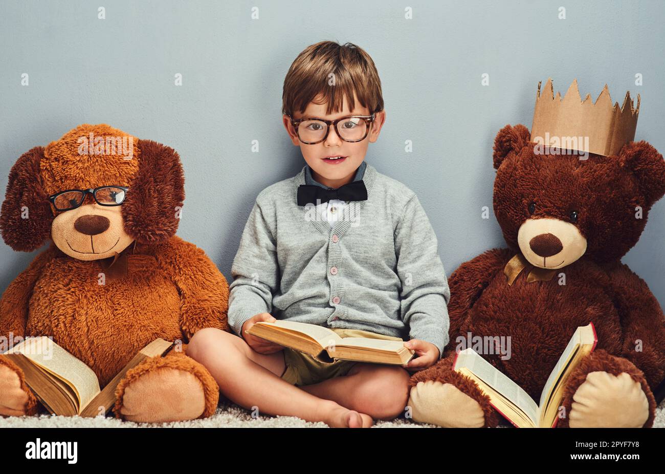 Tutti leggiamo insieme. Studio ritratto di un ragazzino intelligente che legge un libro accanto ai suoi orsacchiotti su uno sfondo grigio. Foto Stock