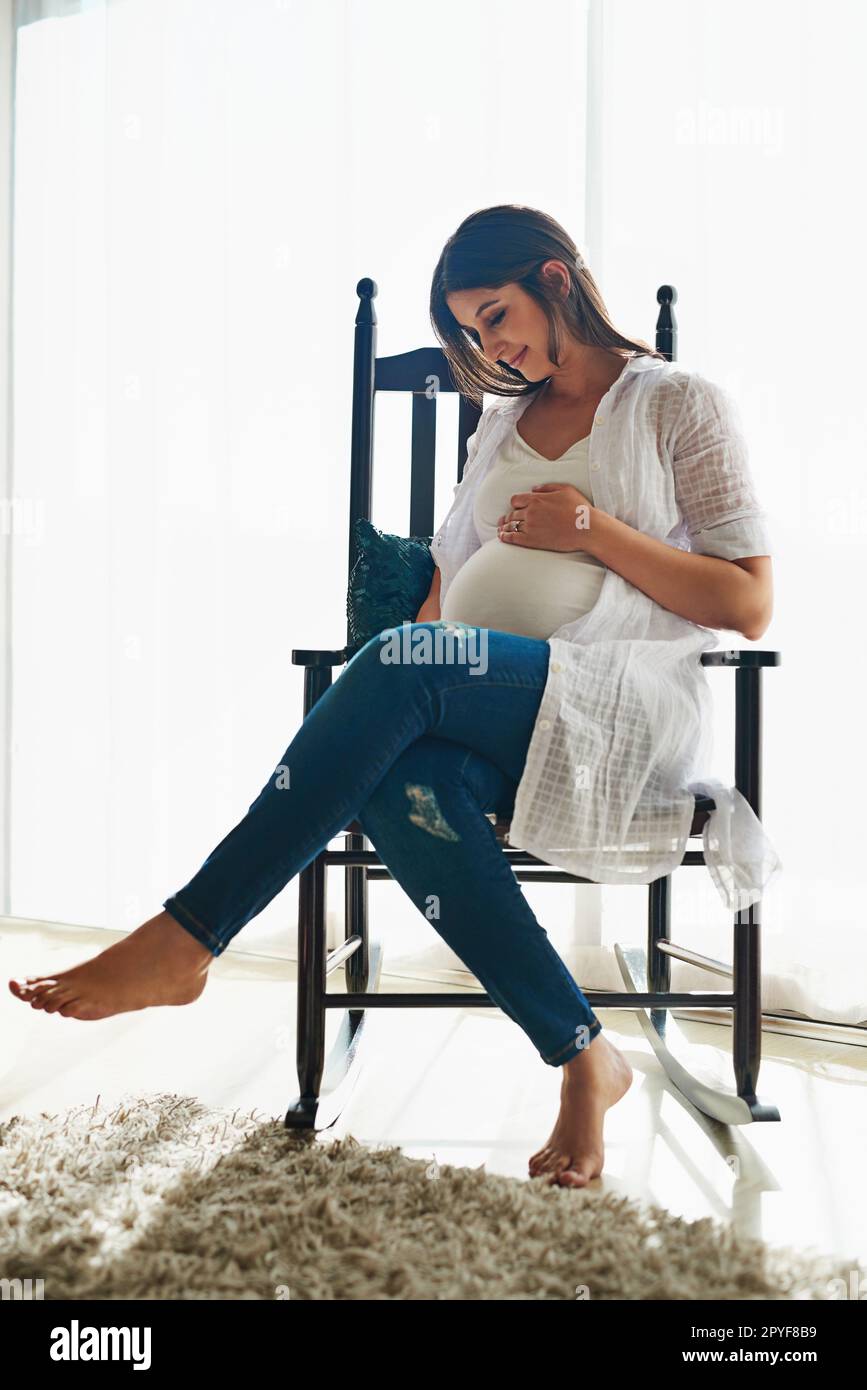 Non vedo l'ora di incontrarti. una donna incinta felice che cullerà il suo ventre mentre si rilassa in una sedia a casa. Foto Stock