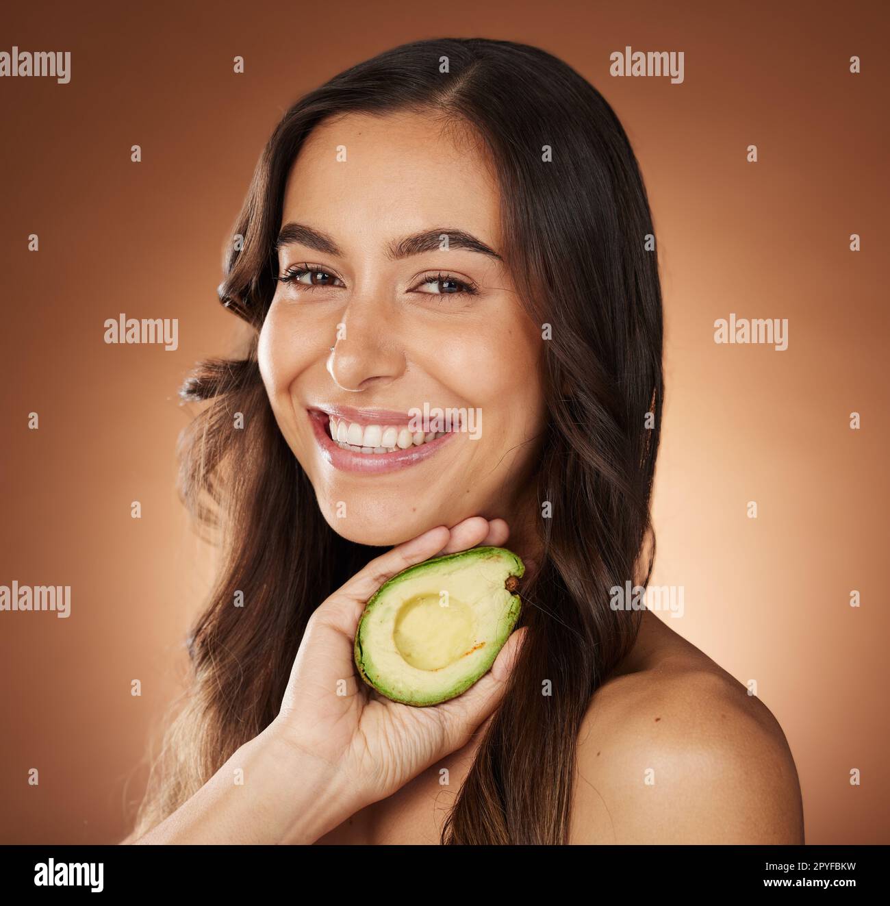 Ritratto di donna, viso e sorriso con avocado per la cura della pelle, dermatologia naturale organica o benessere cosmetico su sfondo marrone studio. Modello, disintossicazione e nutrizione per la cura del viso di lusso Foto Stock