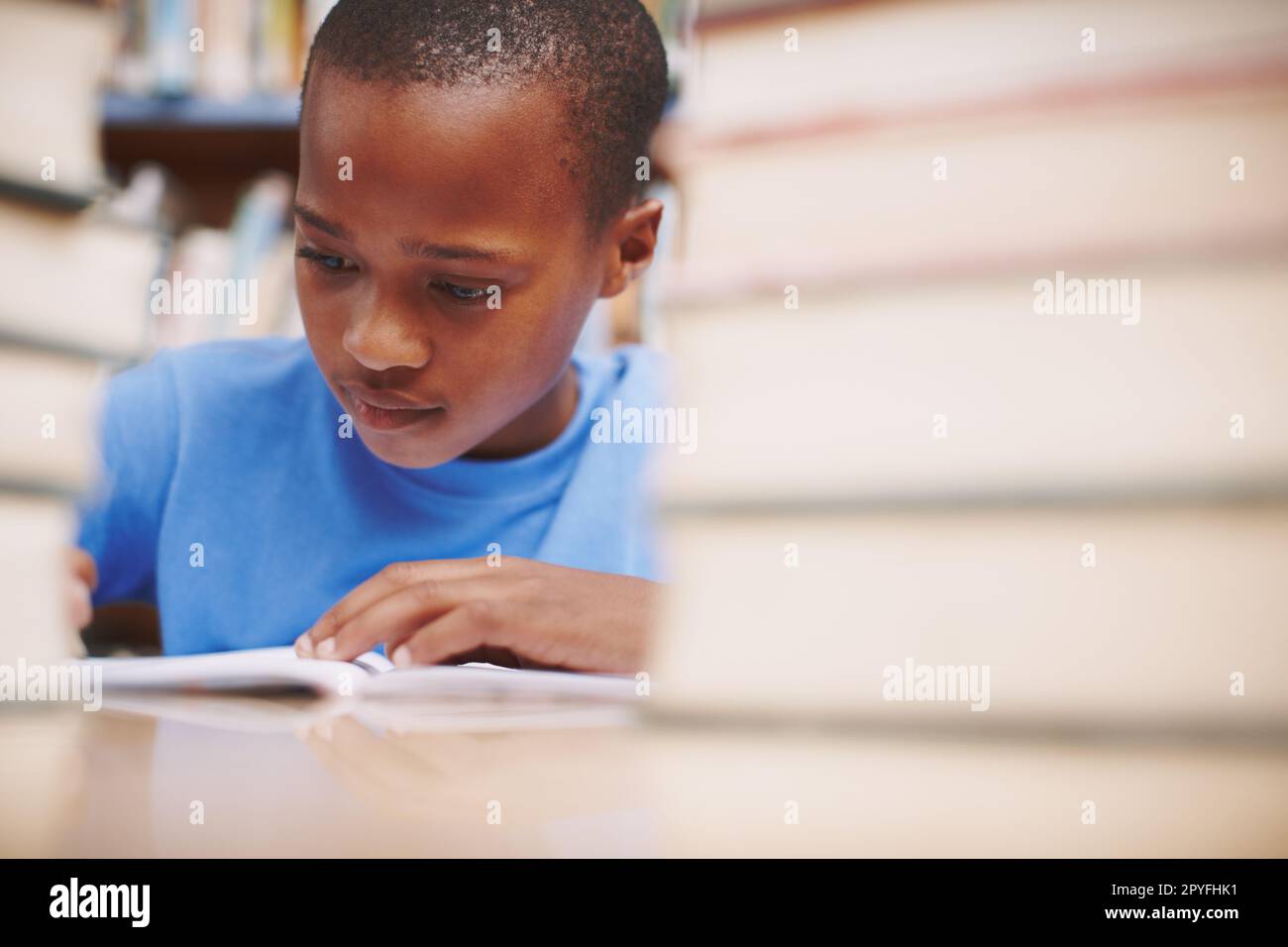 Dedicando tutta la sua attenzione ai libri. Un giovane ragazzo carino che scrive una storia in biblioteca mentre circondato da libri. Foto Stock