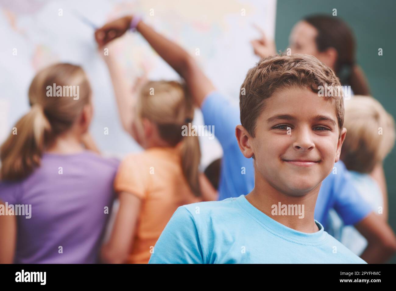 Il mondo ti aspetta. Un ragazzo simpatico che ti sorride mentre i suoi compagni di classe e l'insegnante guardano una mappa del mondo sullo sfondo. Foto Stock