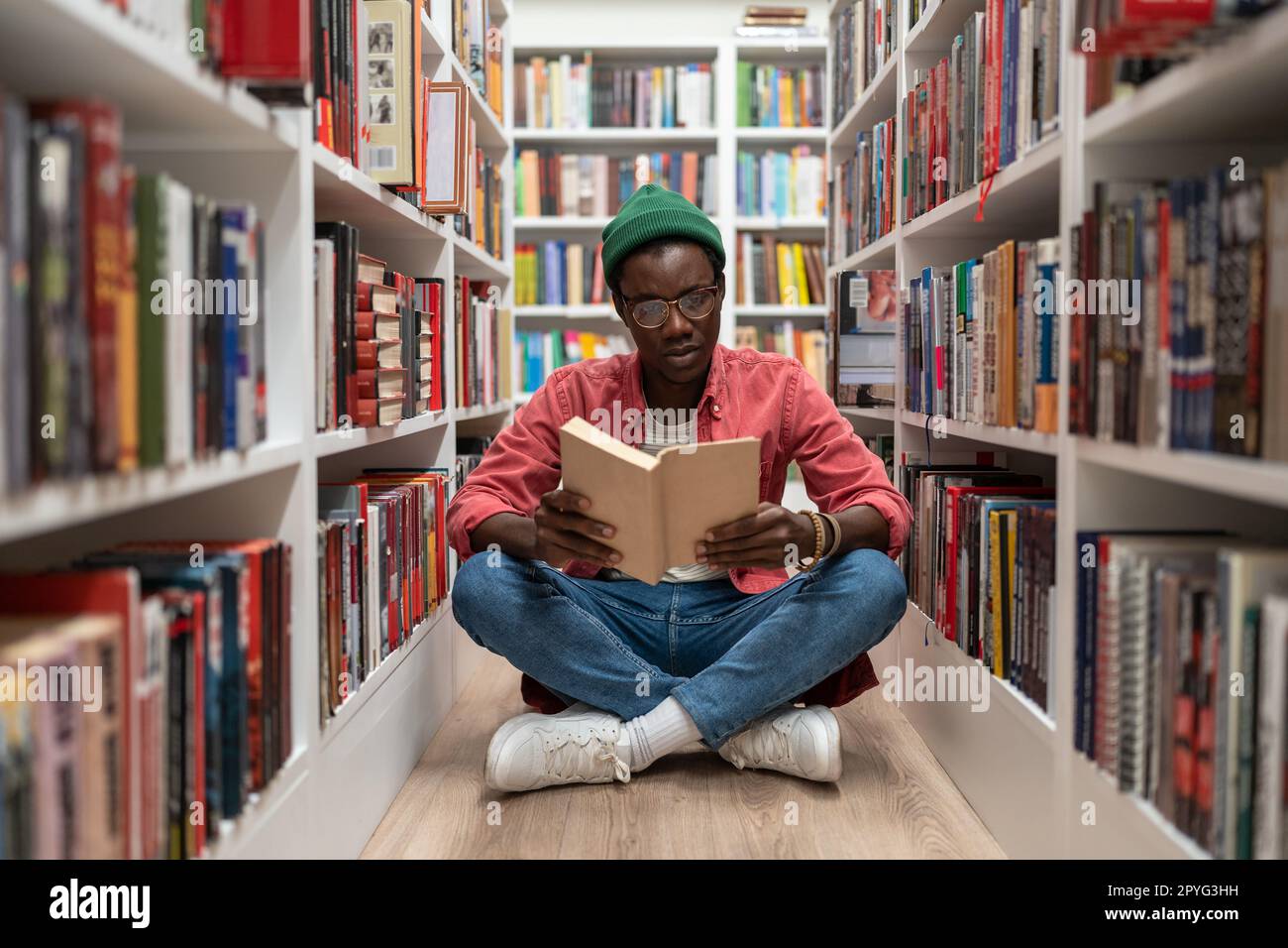 Studente afroamericano uomo lettura libro di ricerca in biblioteca universitaria seduto sul pavimento. Foto Stock