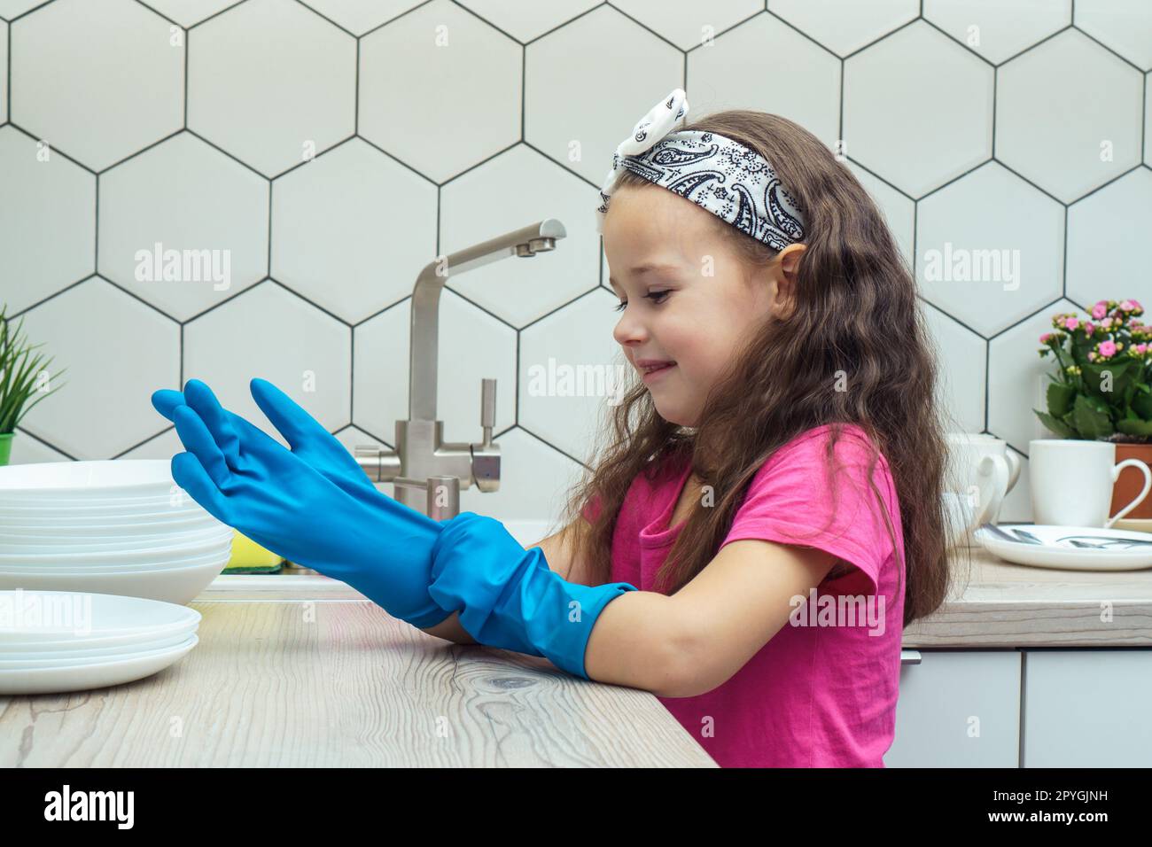 Bambina felice che indossa guanti blu per lavare il piatto nel lavandino della cucina. Concetto di pulizia domestica. Foto Stock