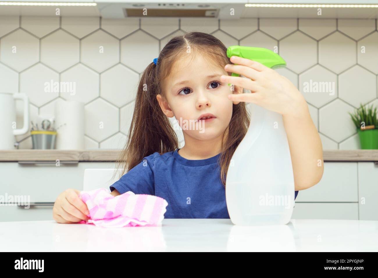 Una bella bambina spinge la leva dello spruzzatore del detergente al tavolo. Ritratto di un bambino che riordina la cucina con uno straccio domestico. Foto Stock