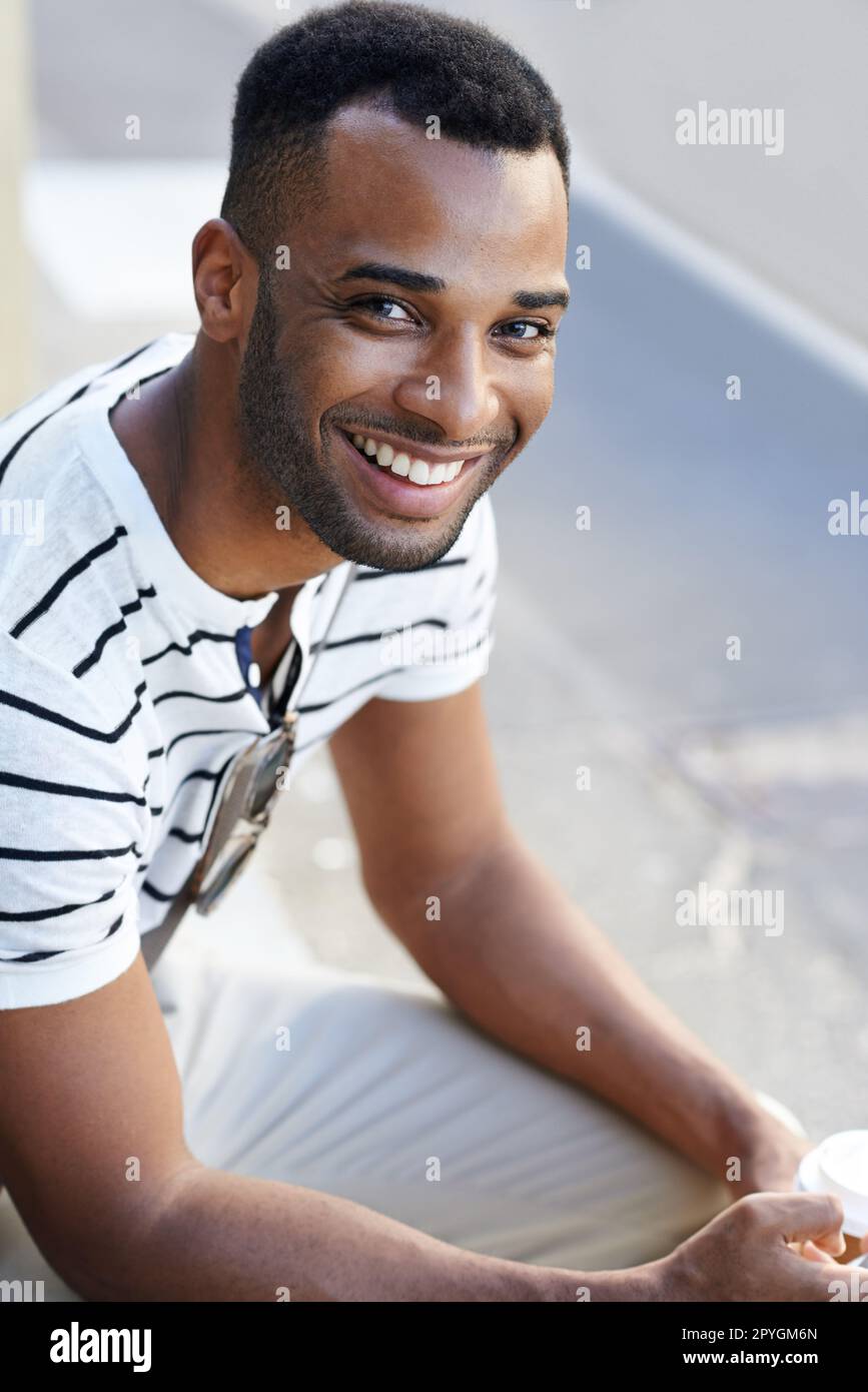 Givng il suo sorriso più luminoso. Un bell'uomo d'affari afro-americano vestito casualmente seduto in città mentre beve un caffè. Foto Stock