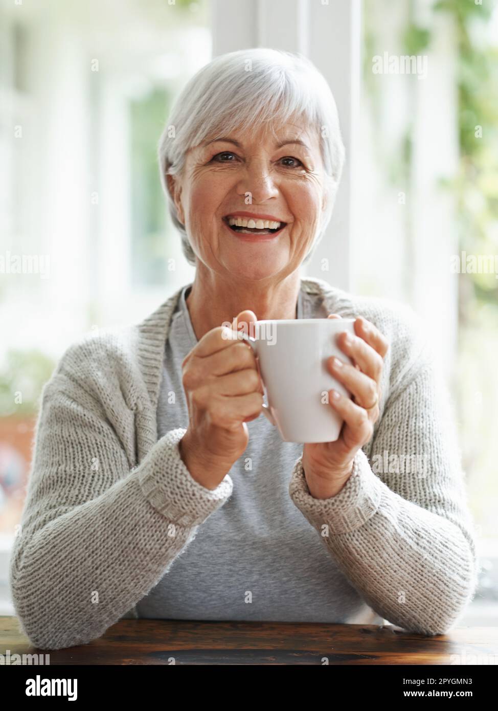 Quella prima tazza di caffè illumina sempre la mia giornata. Una donna anziana felice godendo una tazza di caffè al suo tavolo da cucina - ritratto. Foto Stock
