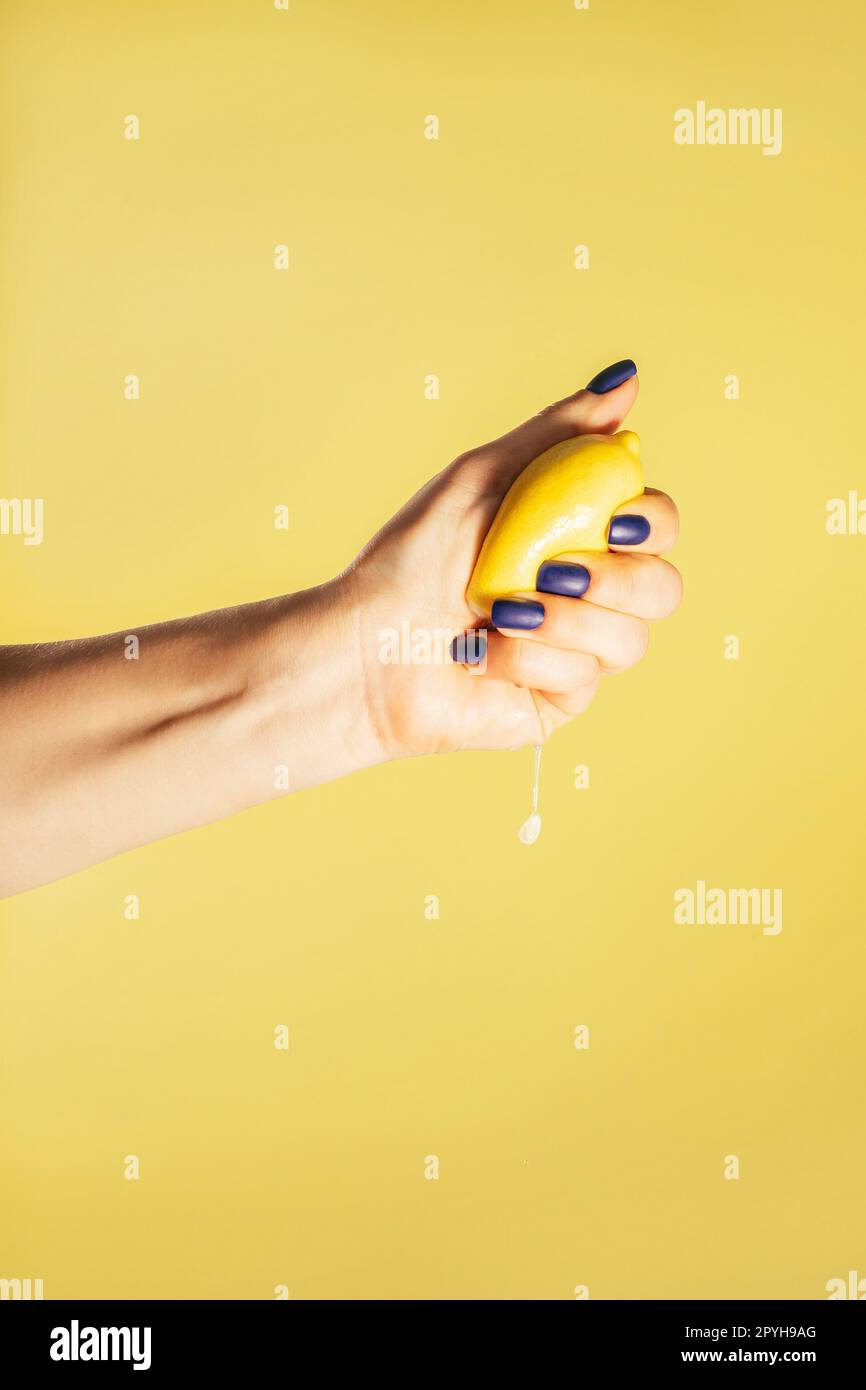 La mano femminile con unghie blu tiene il limone fresco isolato sul giallo. Spremere il succo. Schiacciamento delle dita. Verticale, spazio di copia Foto Stock