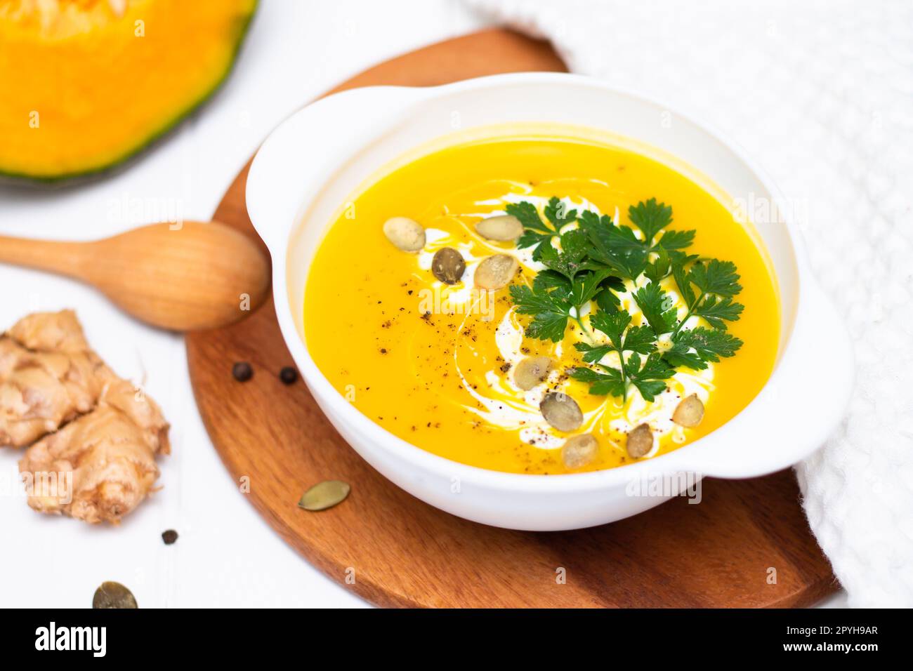 Un piatto di zuppa di crema di zucca gialla con crema, erbe e semi su un tavolo bianco vicino alla zucca cruda e un cucchiaio di legno. Foto Stock