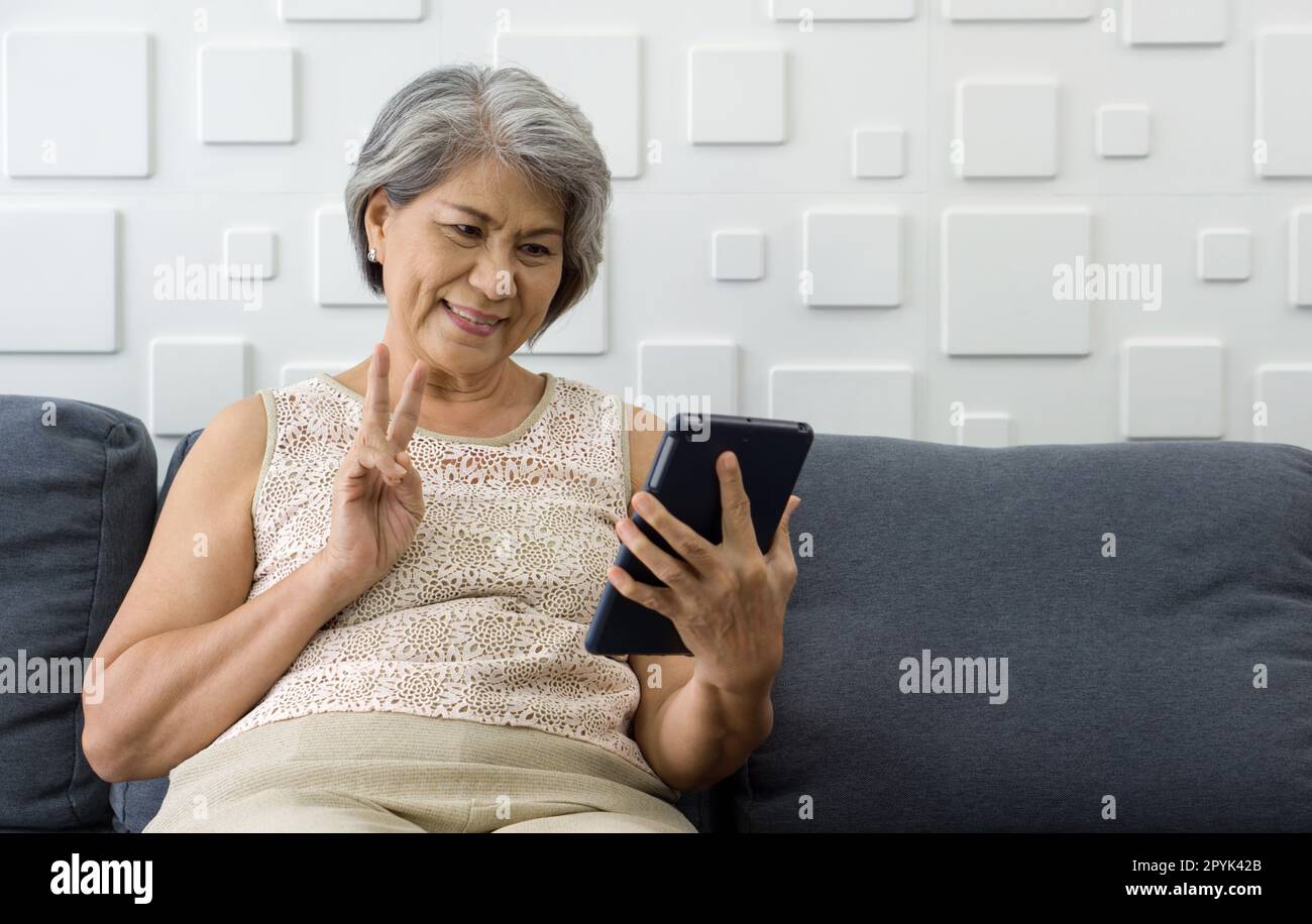 La vecchia donna dei capelli grigia asiatica mostra il segno V tramite la comunicazione remota attraverso il monitor del computer del tablet mentre siede sul divano nel soggiorno. Foto Stock