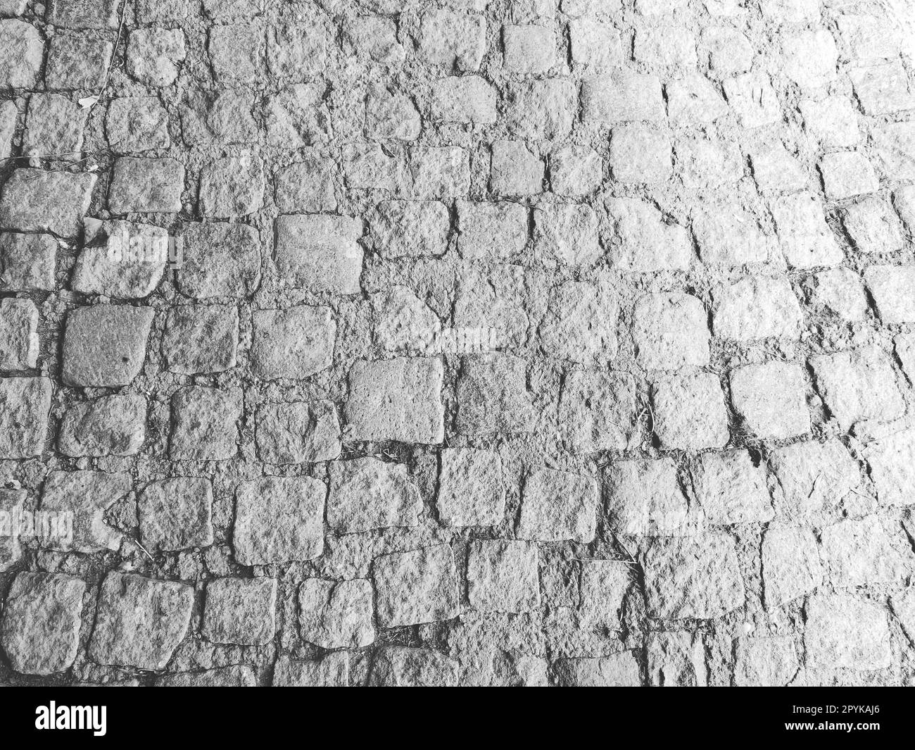 Pavimentazione pavimentata con pietre naturali irregolari. Tra i ciottoli frantumati ci sono piccoli ciottoli, terra, sabbia, erba. Nella vecchia città europea. Fotografia orizzontale monocromatica in bianco e nero Foto Stock