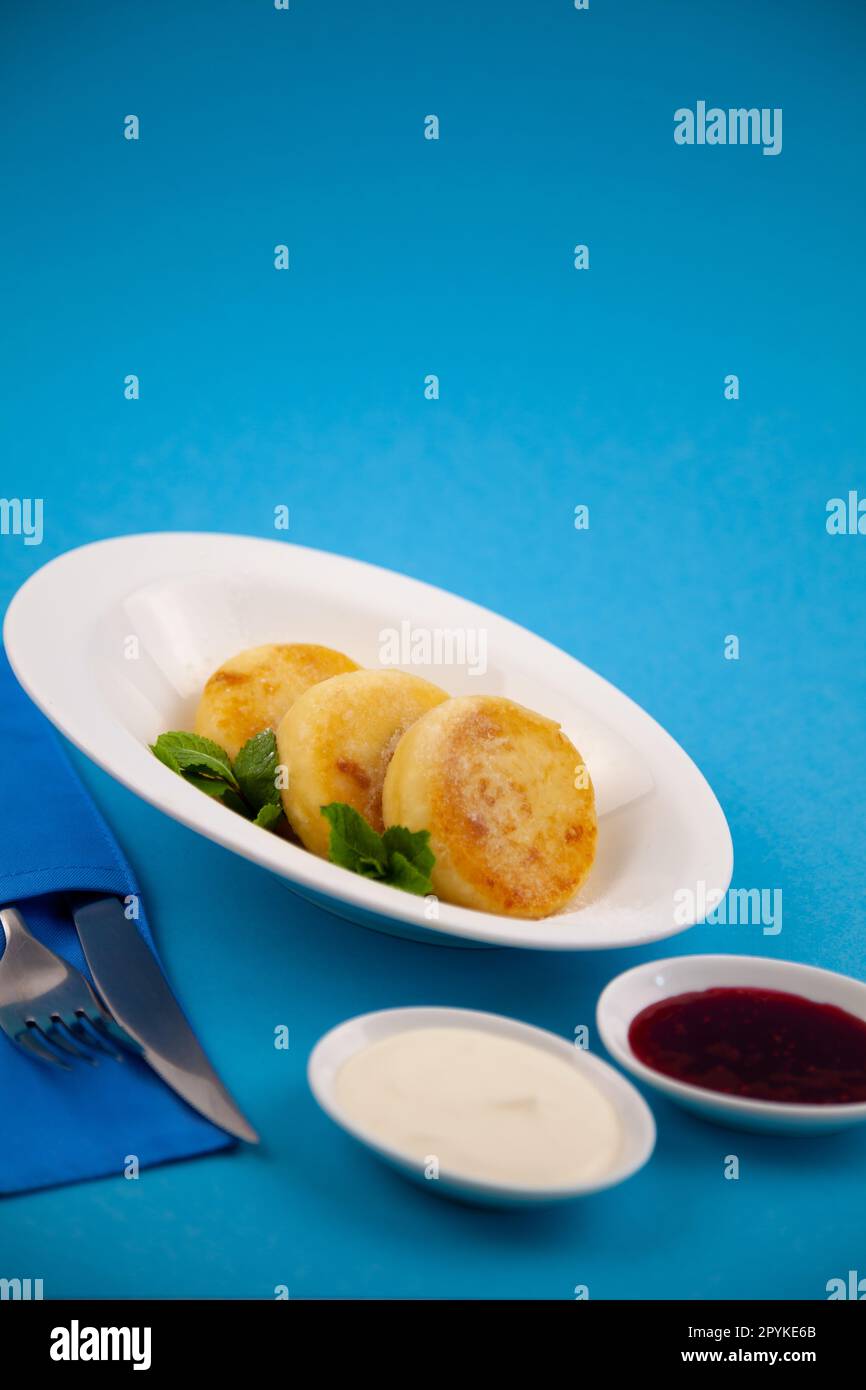 Frittelle di formaggio ricotta rotonda in un piatto bianco su sfondo blu. Cibo prima colazione. Cibo sano. Salsa di panna acida e menta. Primo piano vista dall'alto. Pasto dessert. Frittelle di ricotta al formaggio ricotta. Foto Stock