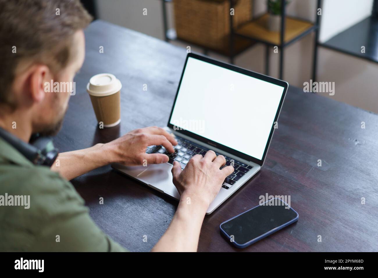 uomo che lavora su un computer portatile in un ufficio loft. uomo seduto alla scrivania con un computer portatile, un telefono cellulare e una tazza di carta da caffè. configurazione tipica dell'area di lavoro, che offre agli spettatori la sensazione di un ambiente di lavoro moderno. Foto di alta qualità Foto Stock