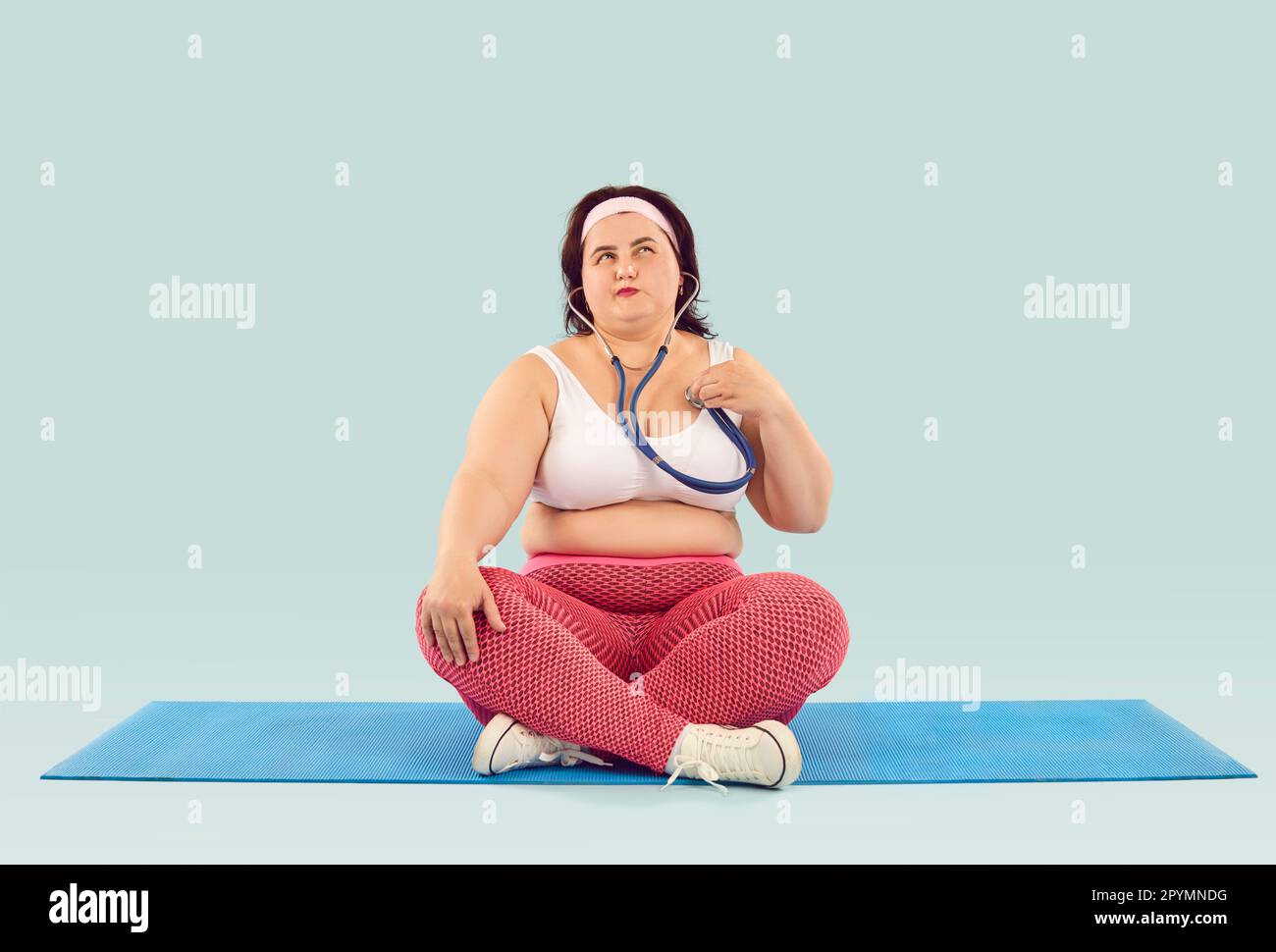 Donna obesa con stetoscopio seduto sul materassino sportivo e pensando ai rischi di malattie cardiache Foto Stock