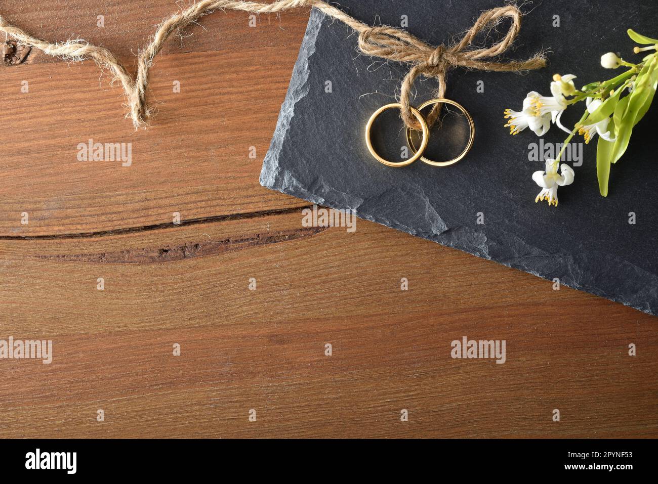 Particolare di due anelli d'oro Uniti con corda di iuta su pietra ardesia su un tavolo di legno rustico e fiori. Vista dall'alto. Foto Stock