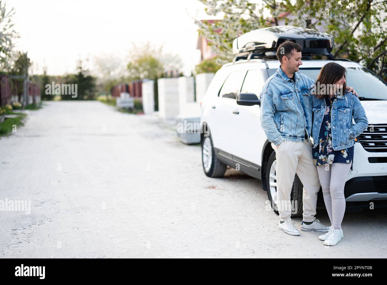 Coppia elegante in giacca jeans in piedi vicino bianco american suv auto con box portabagagli sul tetto. Foto Stock