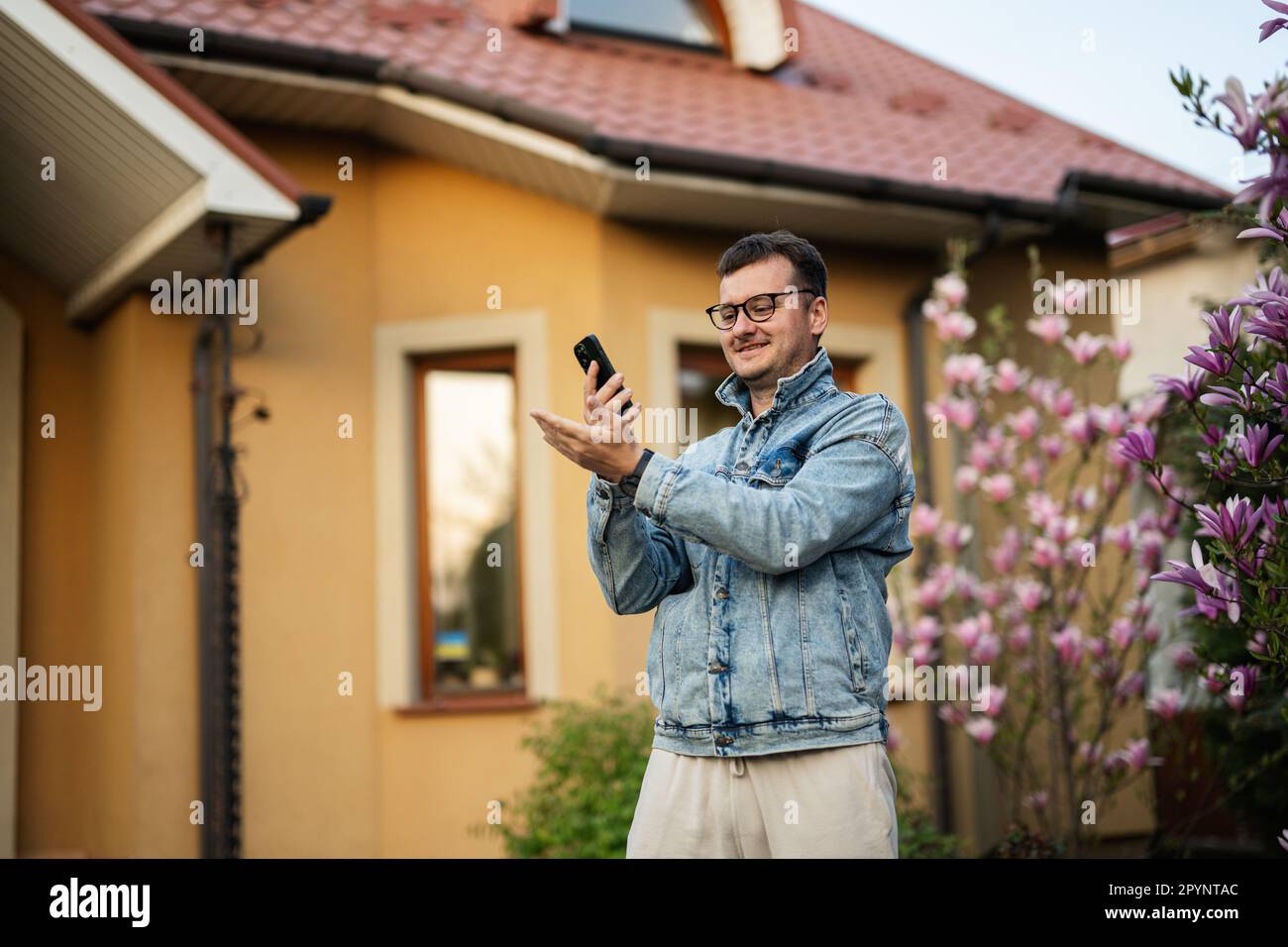 Ritratto di un uomo elegante in occhiali e giacca di jeans, con il telefono cellulare posato contro la sua casa. Foto Stock