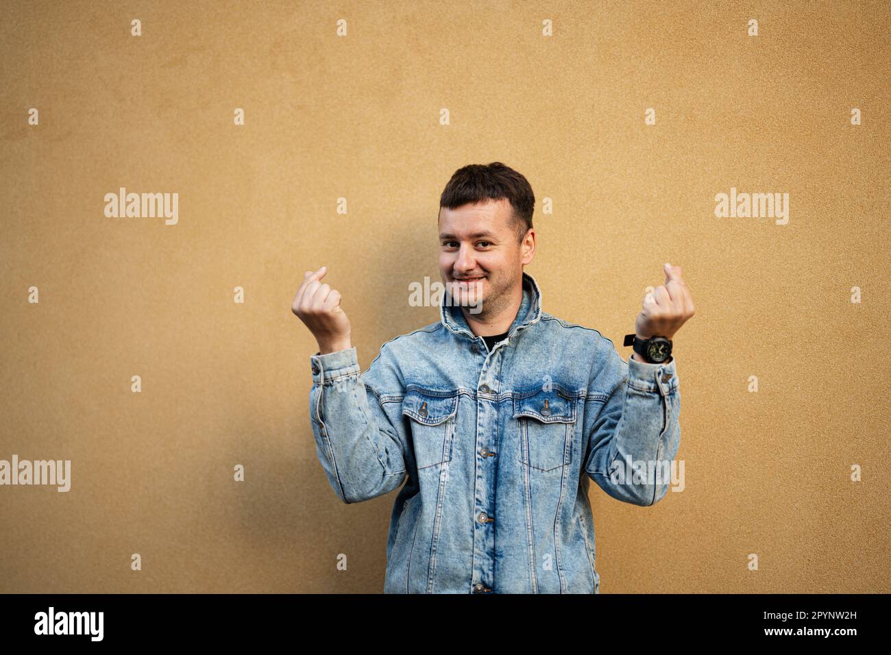 Il ritratto di un uomo elegante in giacca jeans contro la parete gialla rende coreano come segno di cuore da dita. Foto Stock