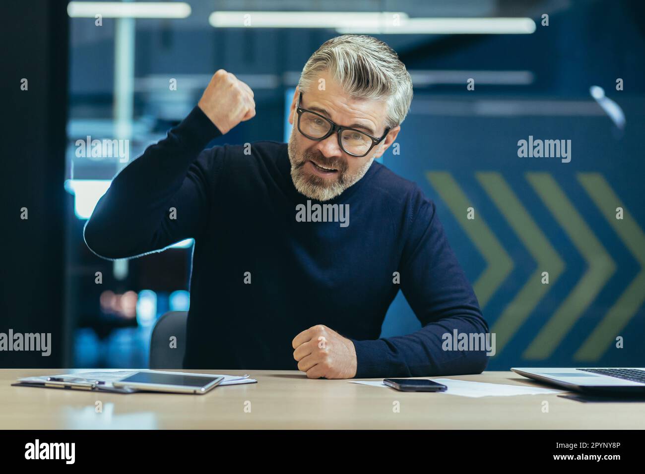 Ritratto di un uomo d'affari anziano arrabbiato seduto in ufficio davanti alla telecamera e gridando, gestendo aggressivamente con le mani, battendo il pugno sul tavolo in modo ordinato. Foto Stock