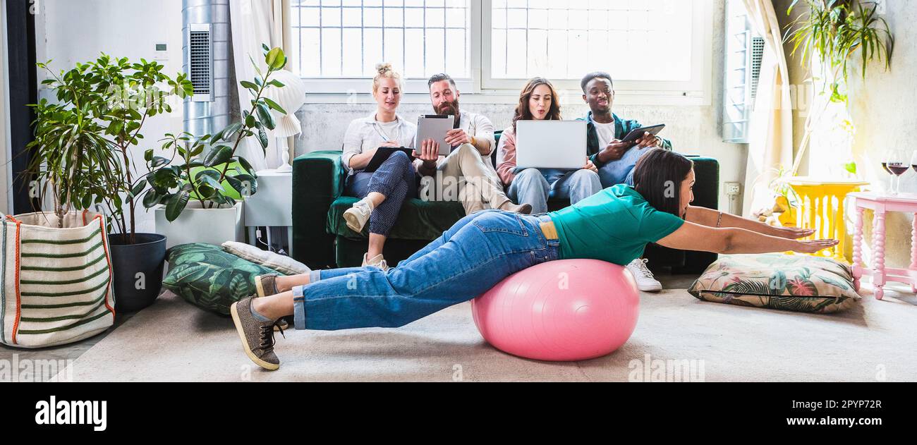 Scena bizzarra e divertente con ragazzi che studiano al computer e una ragazza che gioca pazzo con una palla pilates. Momento divertente durante una pausa per prepararsi per un Foto Stock