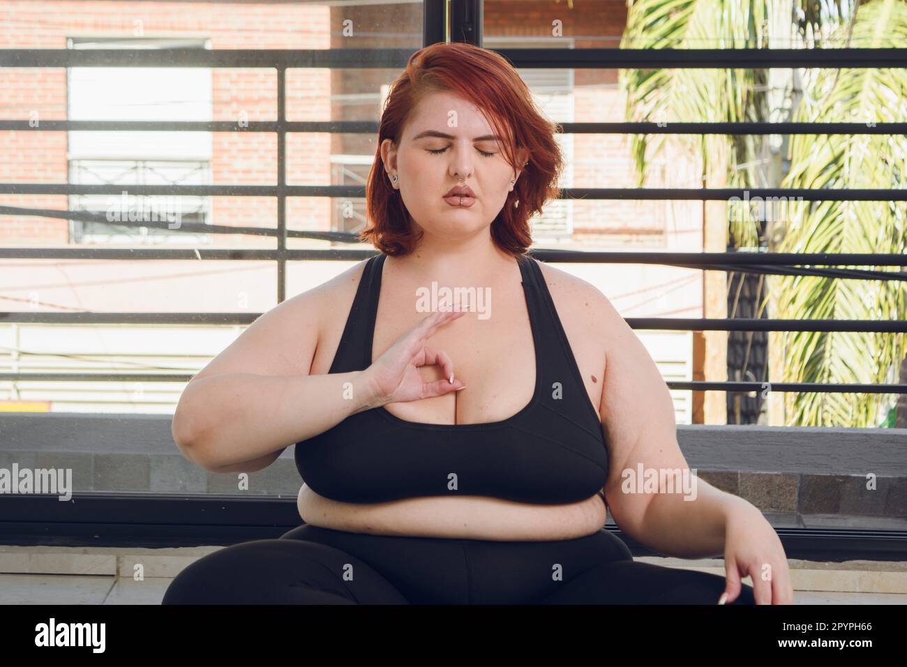 giovane latino più dimensione donna seduta con gli occhi chiusi meditando con mano sul petto in posizione mudra gyan, vista frontale al coperto, yoga concetto. Foto Stock