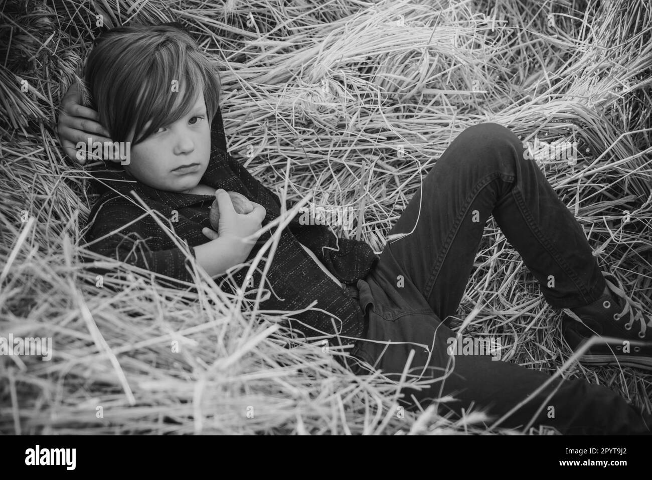 Ragazzo di capretto che tiene la foglia d'oro e si trova sul fieno. Ragazzo dai capelli fieni giace su fondo di fieno e mangia una mela. Ciao estate - Ciao autunno. Capretto su un autunno Foto Stock