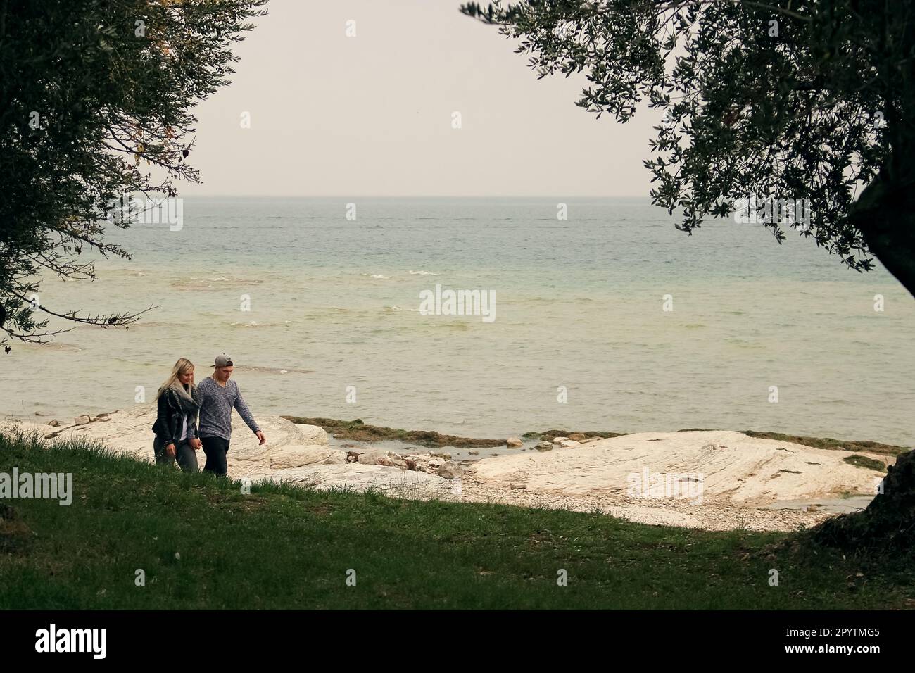 una giovane coppia tradizionale, un uomo bianco e una donna bianca, innamorati e in mano, camminano lungo la riva di un grande lago senza paura della pioggia. Foto Stock