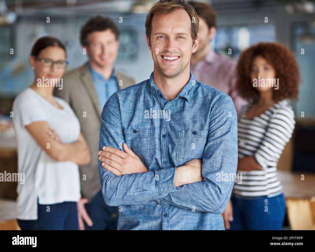 Ambienti di lavoro positivi per team forti. Uomo sorridente con le braccia incrociate e colleghi amichevoli sullo sfondo. Foto Stock