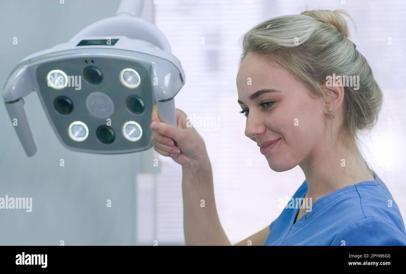 Bella infermiera che regola la luce dentale in clinica. L'assistente dentista prepara l'apparecchiatura di illuminazione prima del trattamento e sorride al paziente Foto Stock