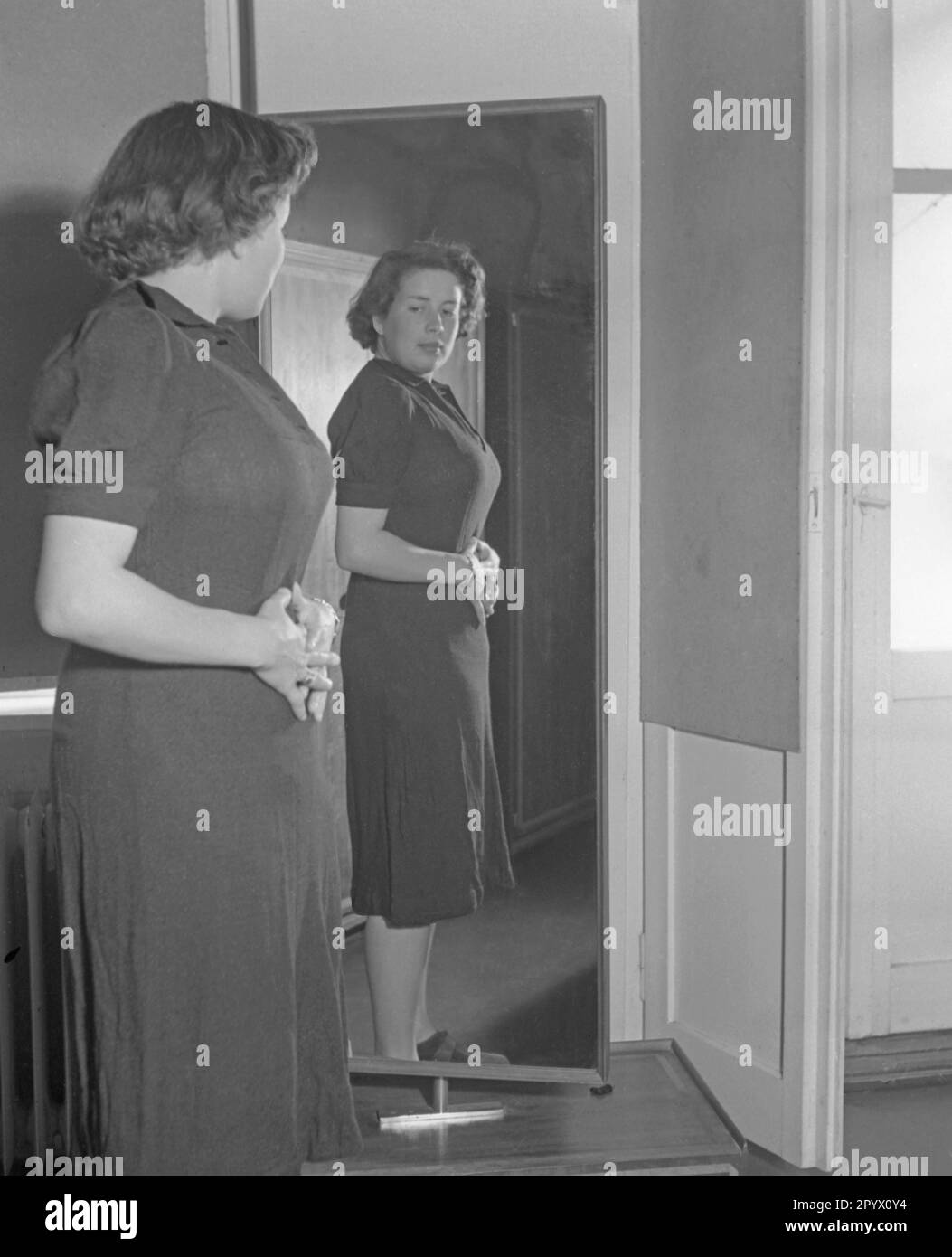 Una donna in un vestito si alza lateralmente di fronte a uno specchio e si guarda mentre le sue mani la coprono il ventre. Foto Stock