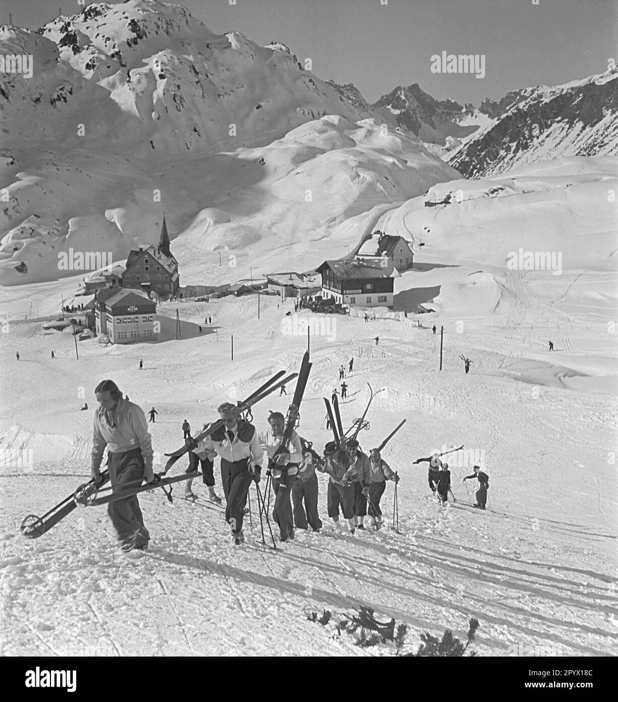 Un gruppo di sciatori sulla strada per la pista nella metà degli anni '1930s. Sullo sfondo il resort St. Christoph con l'ex ospizio, che è utilizzato come hotel dal 20th ° secolo. Foto Stock