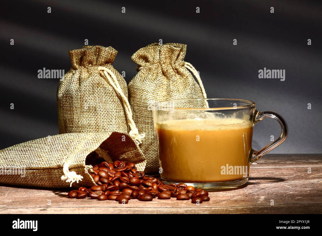 Sacchi di chicchi di caffè crudi tostati e una tazza di caffè con un riflesso di una finestra cieca sullo sfondo Foto Stock