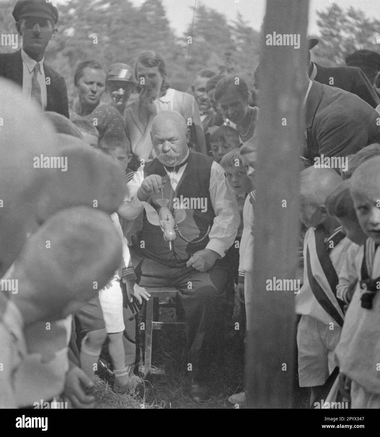 Un uomo mira a qualcosa con un uccello di legno attaccato ad una corda. I bambini intorno a lui stanno guardando il suo approccio (foto non data). Foto Stock