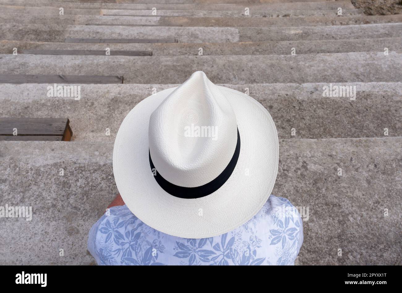 Un uomo che indossa una camicia blu e un grande cappello beige Panama sulla testa, seduto su alcuni gradini di pietra e con la schiena alla macchina fotografica. Cadice, Spagna Foto Stock