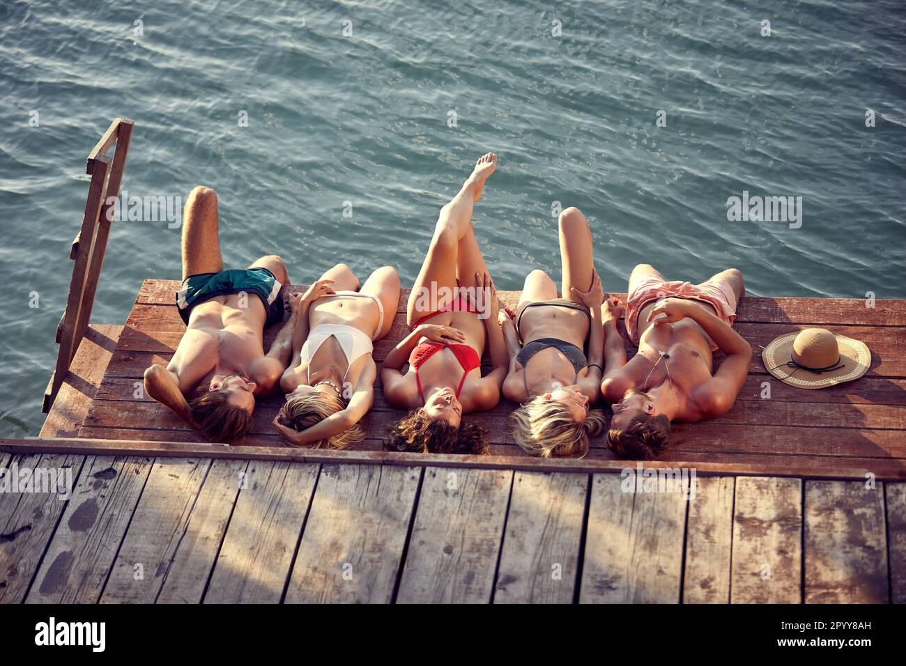 Gruppo di giovani amici adagiato su un molo di legno in acqua, prendendo il sole. Giovani uomini e donne in vacanza. Vacanza, convivenza, concetto di stile di vita. Foto Stock