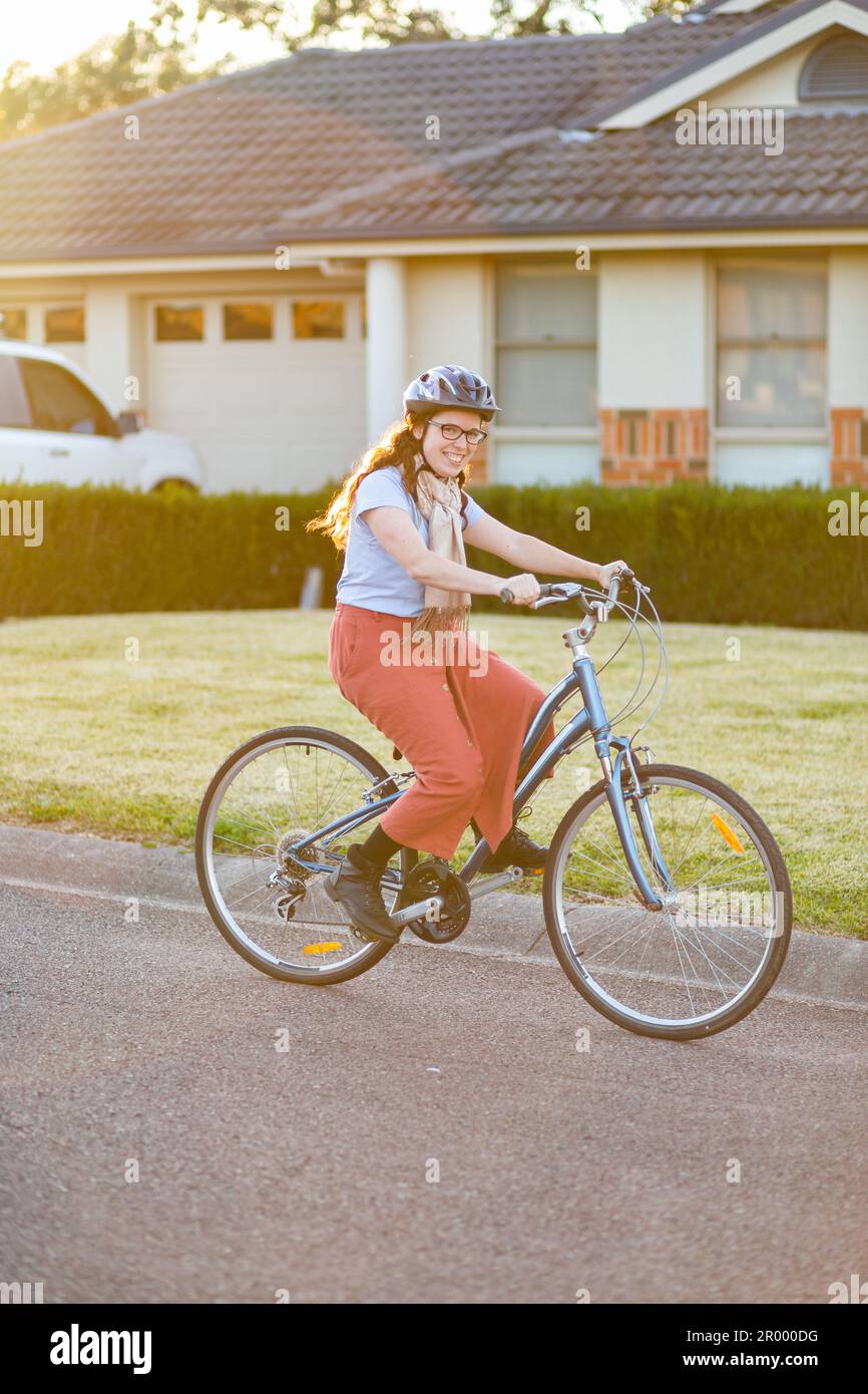 Giovane donna ventenne in sella a una bici su strada suburbana con casco da ciclismo Foto Stock