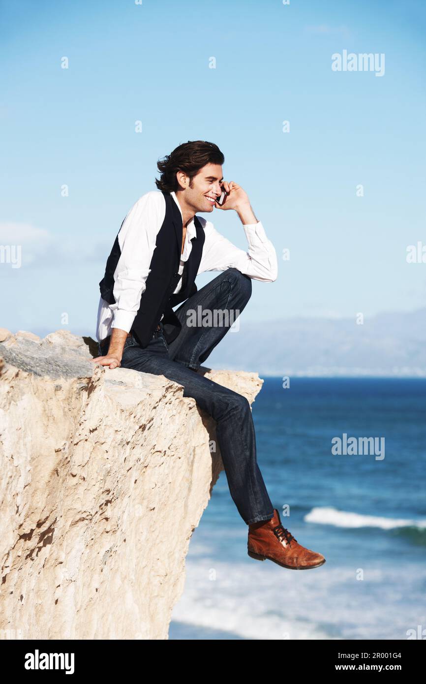 Non indovinate mai da dove si chiama l'IM. Un giovane sorridente che chiacchiera al telefono mentre si siede su una scogliera che si affaccia sull'oceano. Foto Stock