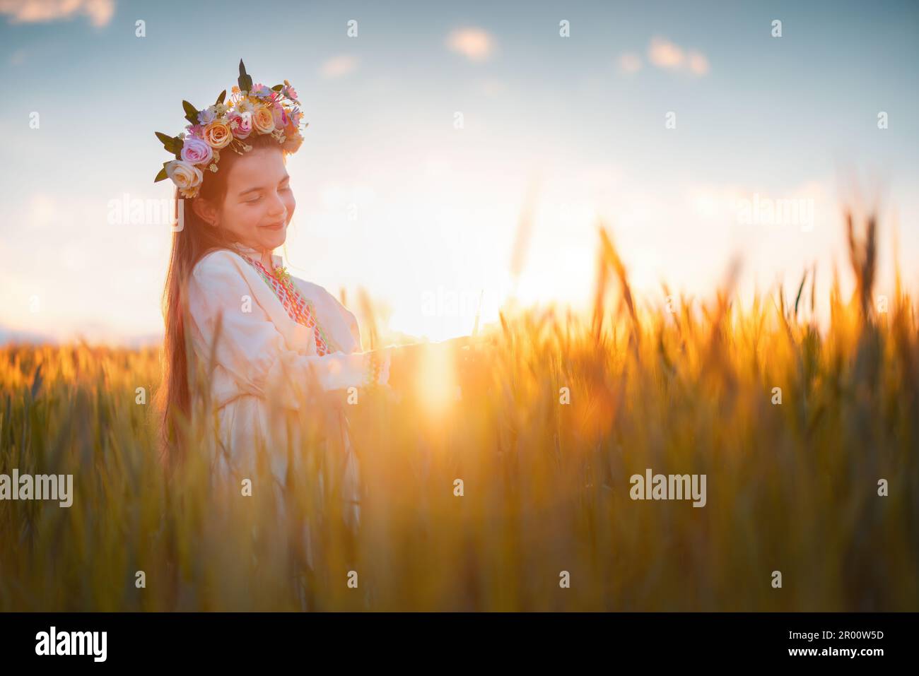 Bella giovane ragazza con cappelletto di fiori, abito folcloristico etnico con ricami tradizionali bulgari durante il tramonto su un campo agricolo di grano Foto Stock