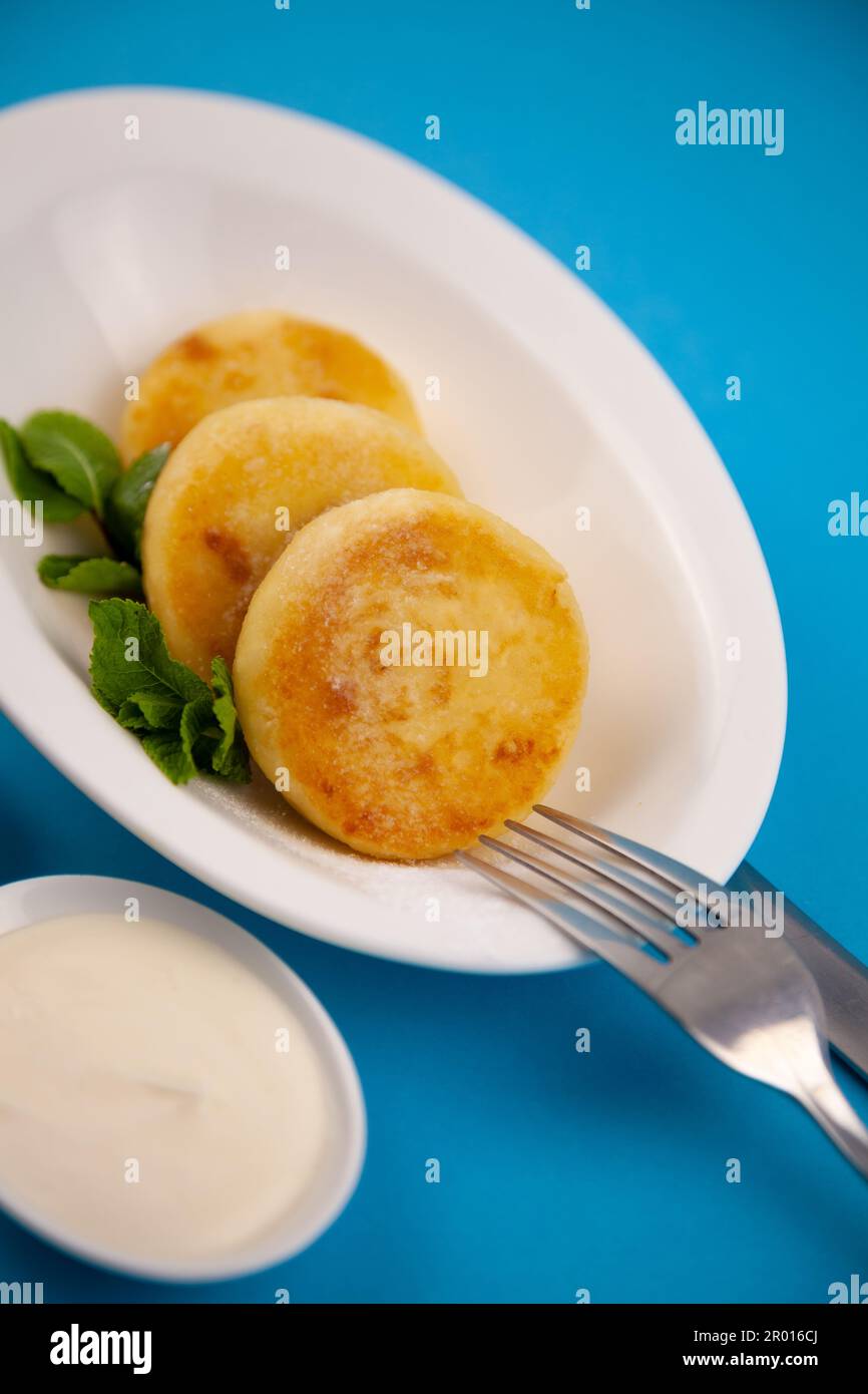 Frittelle di formaggio ricotta rotonda in un piatto bianco su sfondo blu. Cibo prima colazione. Cibo sano. Salsa di panna acida e menta. Primo piano vista dall'alto. Pasto dessert. Frittelle di ricotta al formaggio ricotta. Foto Stock
