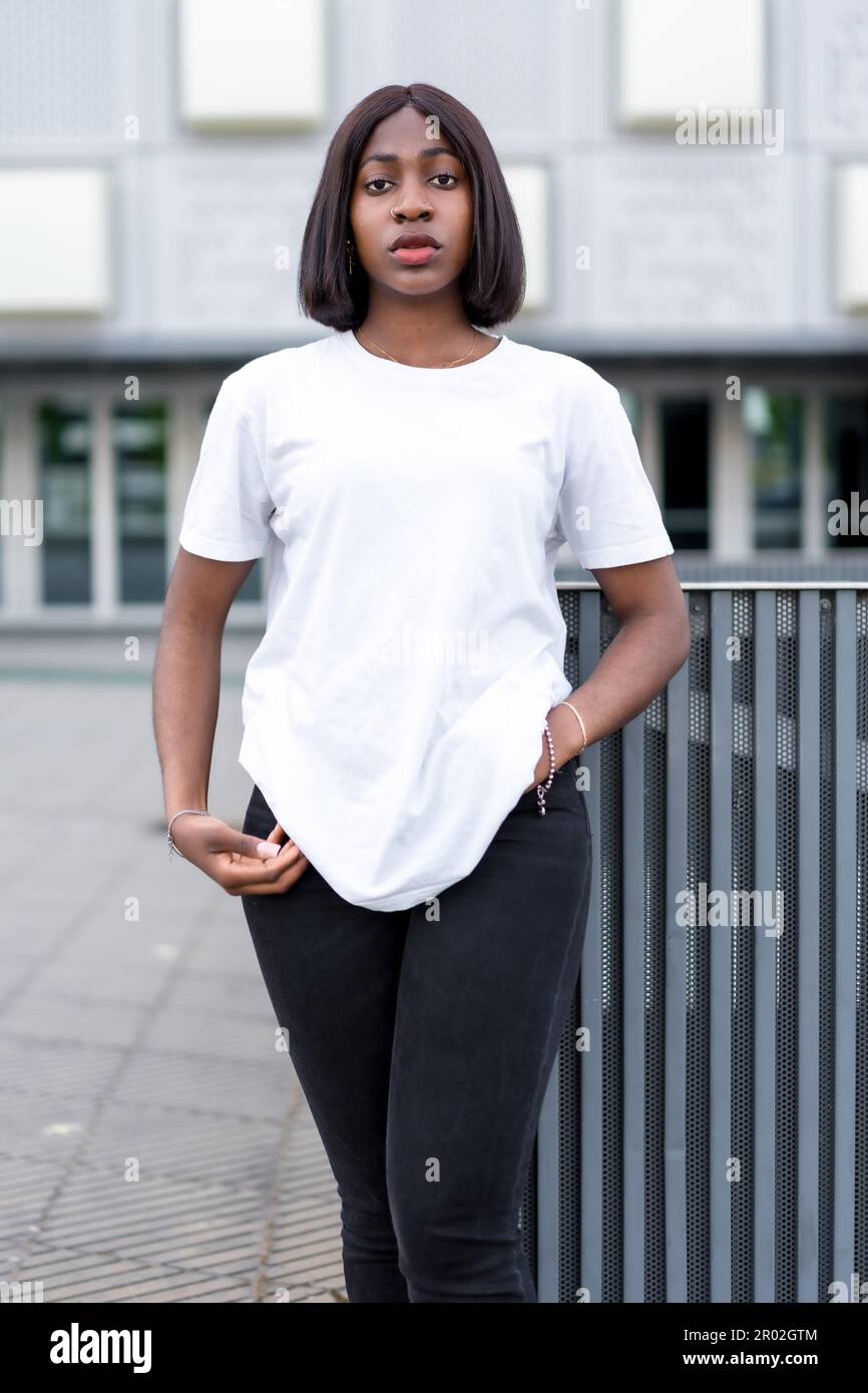 Cool senza sforzo: Un ritratto di una giovane donna nera elegante con un edificio grigio come sfondo, indossando una t-shirt bianca e pantaloni neri Foto Stock