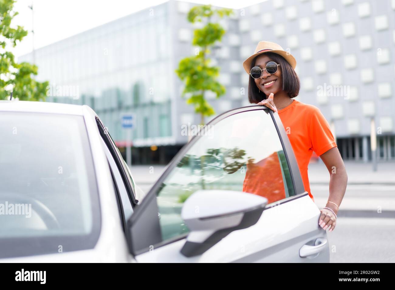 Posa con stile: Donna nera che indossa una camicia arancione e occhiali da sole che aprono la porta dell'auto Foto Stock