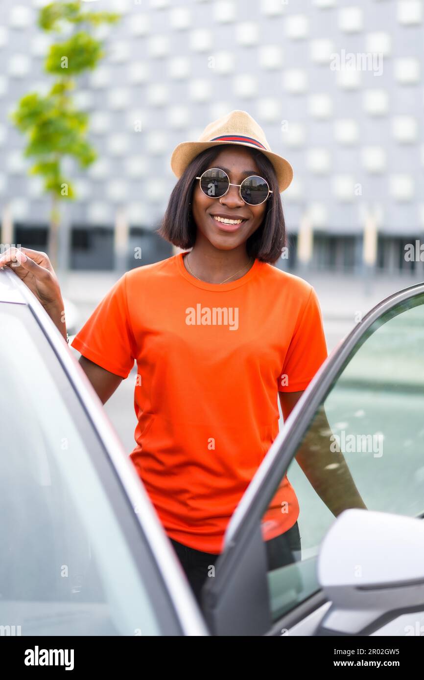 Donna nera con occhiali da sole che aprono la porta dell auto indossa una camicia estiva arancione e sorride in citta Foto stock Alamy