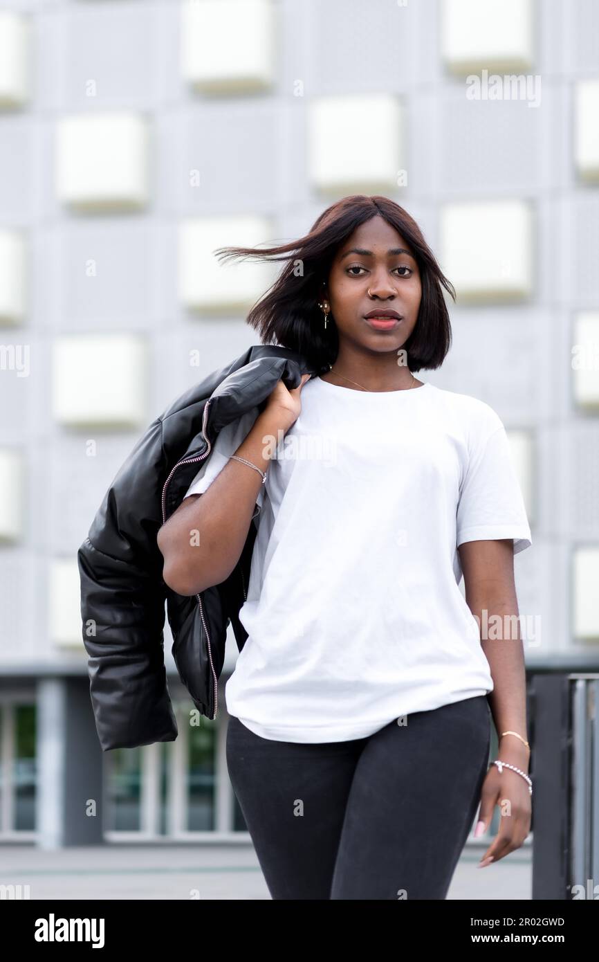 Città alla moda: Una giovane donna nera posa in un elegante tee bianco e pantaloni neri, con un edificio grigio come sfondo urbano chic Foto Stock