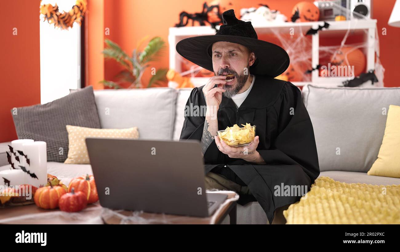 Giovane uomo calvo che indossa costume da mago mangiare patatine patate guardando il film a casa Foto Stock